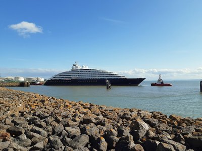 Das Traumschiff Scenic Eclipse II liegt im Hafen. Im Vordergrund liegen Steine und der Himmel ist blau.