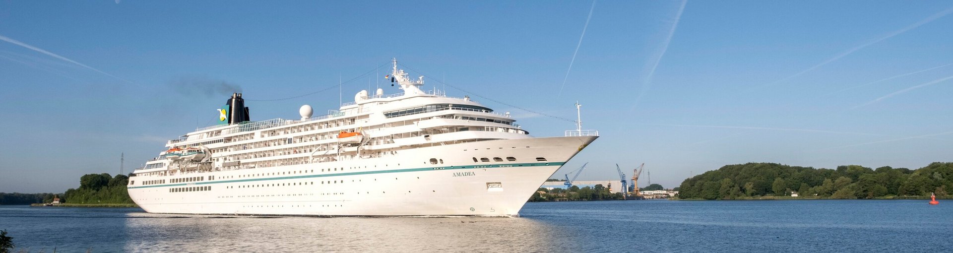Ein weißes Kreutzfahrtschiff fährt auf den Nord-Ostsee-Kanal entlang. Der Himmel ist blau. 