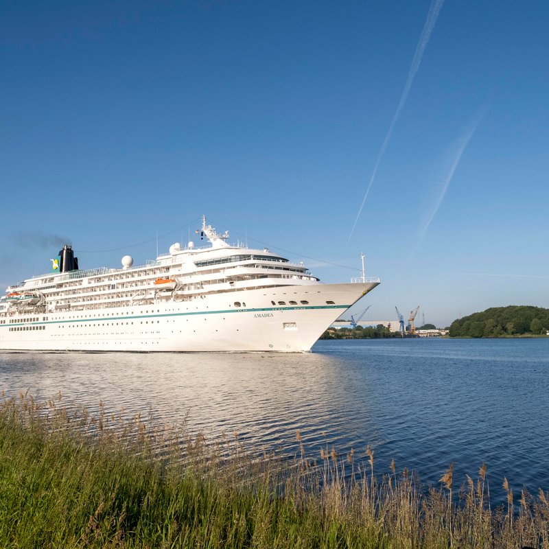 Ein weißes Kreutzfahrtschiff fährt auf den Nord-Ostsee-Kanal entlang. Der Himmel ist blau. 